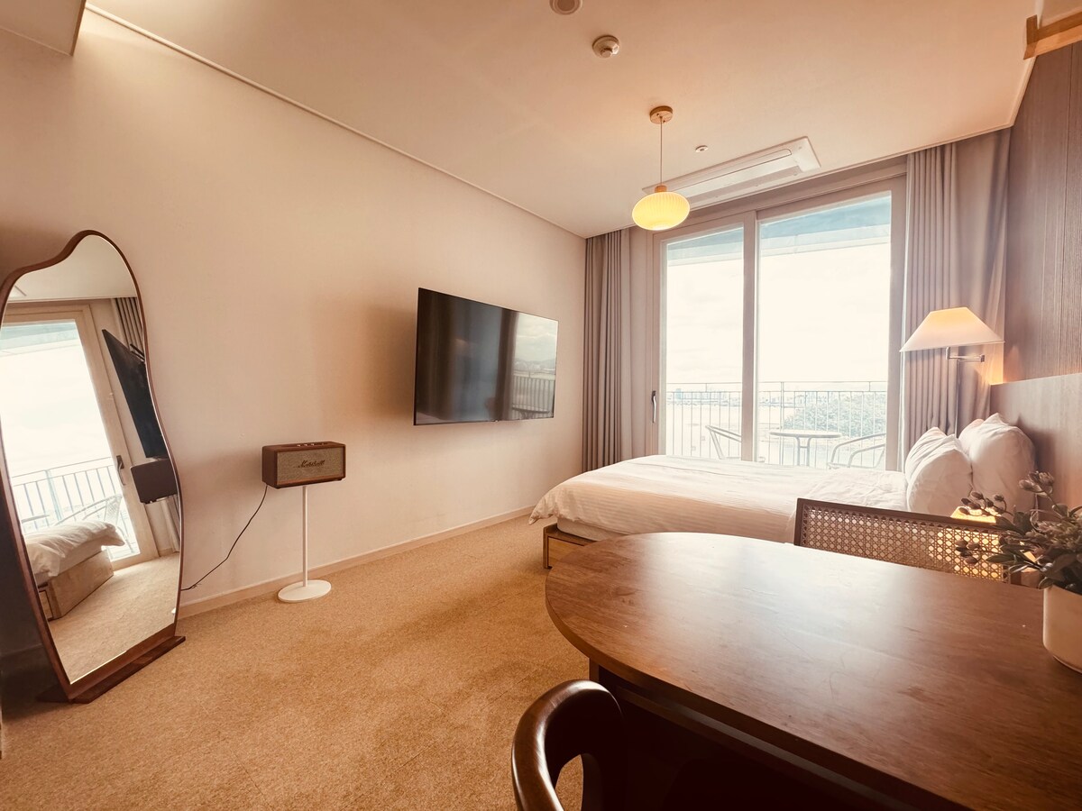 A modern room features a spacious bed against a wall with a large window providing natural light. A sleek mirror stands nearby, and a flat-screen TV is mounted for entertainment. A small desk is positioned next to a stylish chair, enhancing the room's functionality.