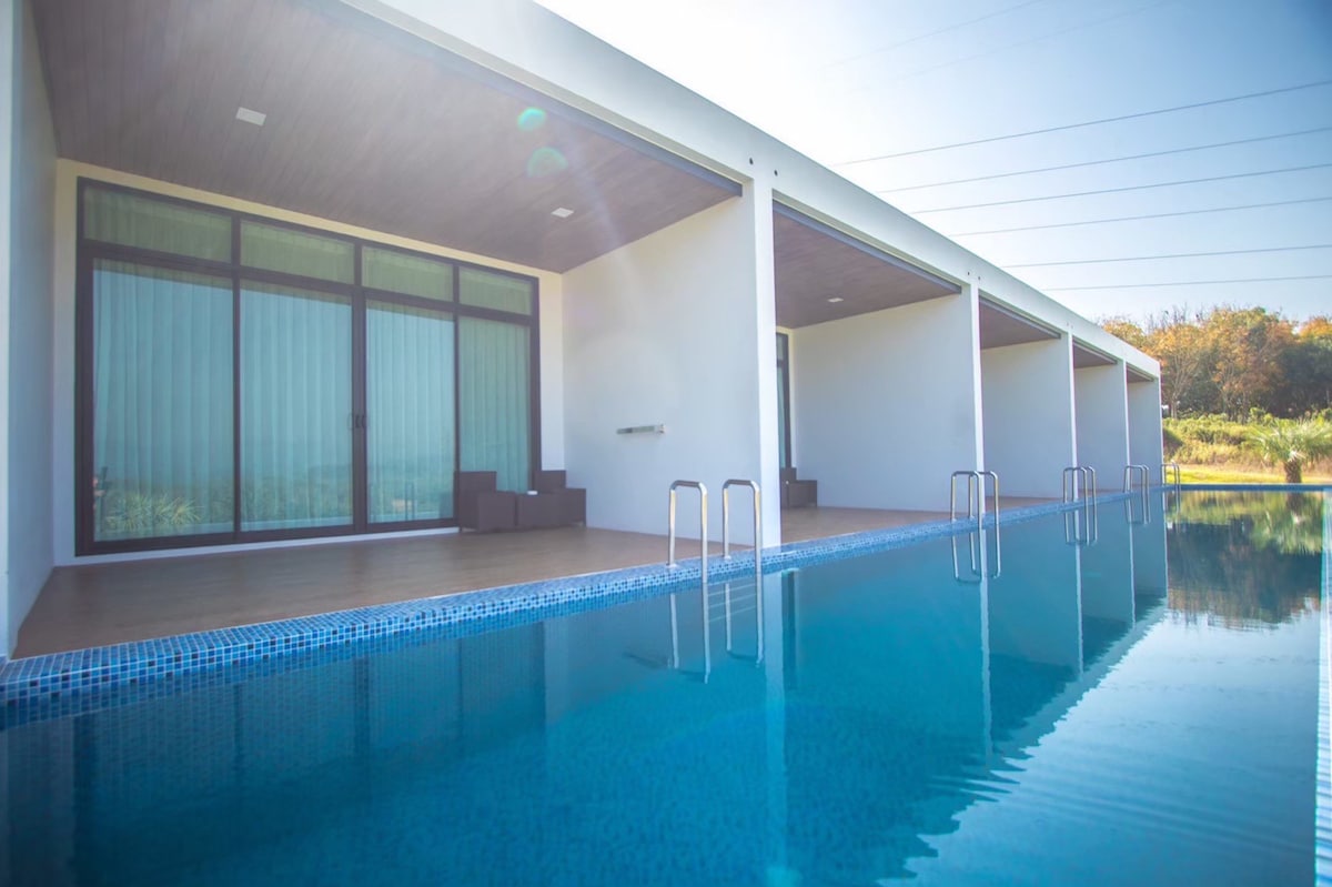A clear swimming pool reflects the adjacent modern structure, which features spacious terraces with dark seating arrangements. Expansive glass doors offer seamless access to the outdoors, allowing natural light to enhance the serene atmosphere.