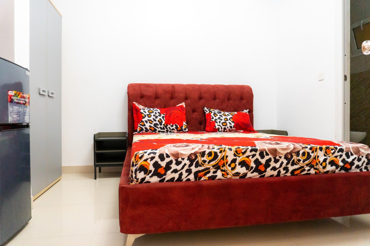 A spacious bed with a vibrant red and leopard print coverlet is centered in the room. Decorative pillows in floral patterns are arranged at the head of the bed. A bedside table and a refrigerator are visible, along with a wardrobe and a doorway leading to a bathroom.