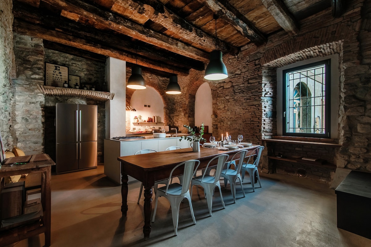 The kitchen and dining area feature exposed wooden beams and rustic stone walls. A wooden dining table with metal chairs is positioned under industrial-style pendant lights. Stainless steel appliances and an open layout enhance the space, which is complemented by large windows allowing natural light.