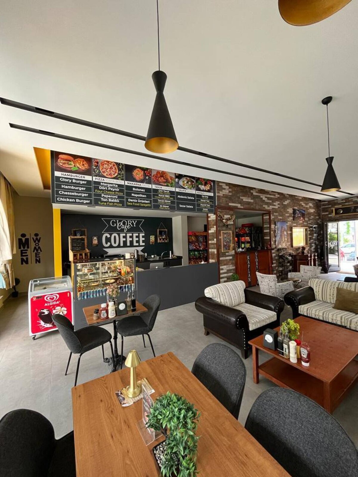 A modern café space features a service counter with a detailed menu displayed above. Comfortable seating areas are arranged with dark and light-toned furniture. Refrigerated displays hold various drink options, and tables are set with decorative plants, creating an inviting setting for guests.
