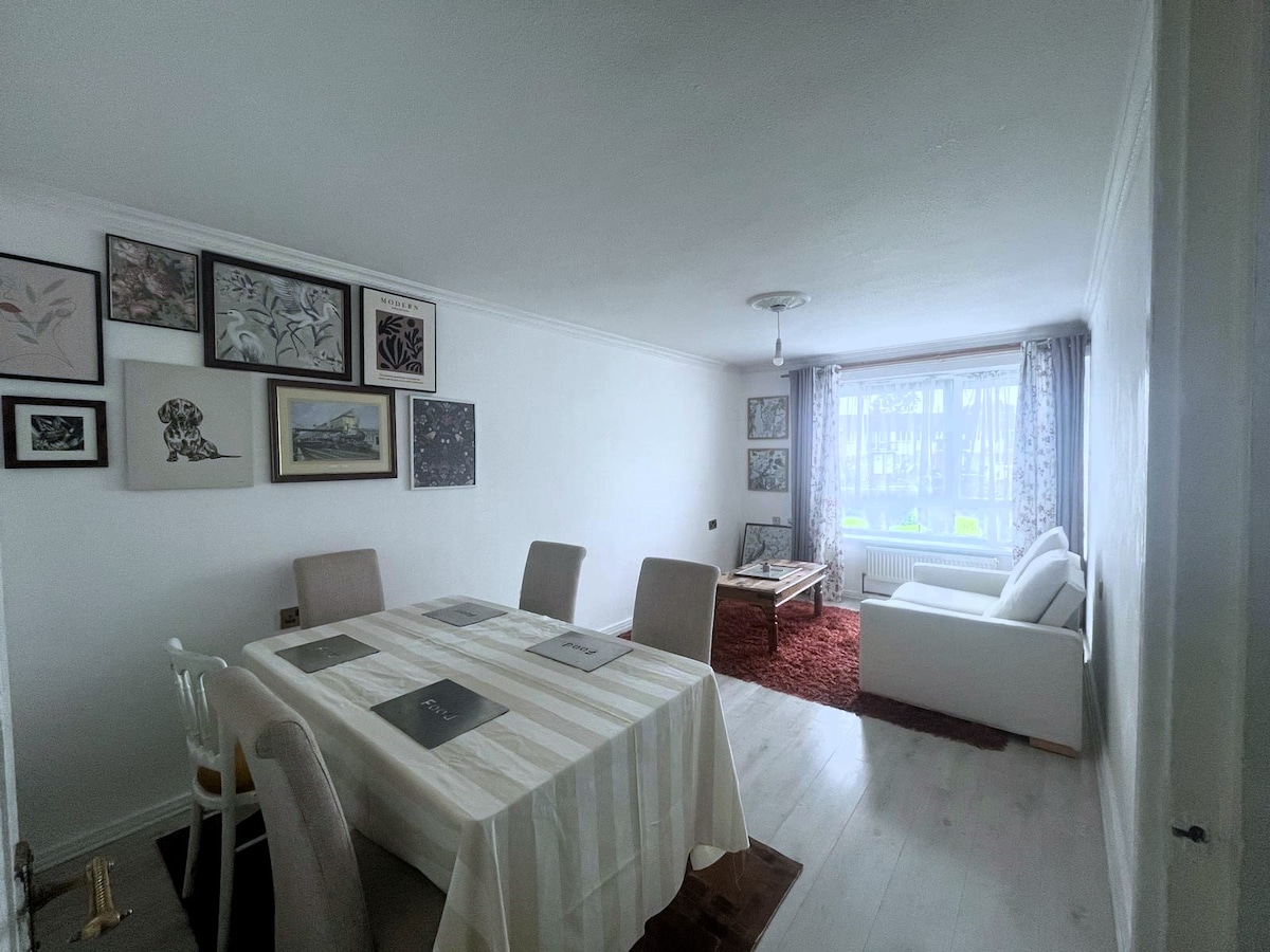 A bright living and dining area features a comfortable white sofa and a rectangular dining table set for six. The walls are adorned with framed artwork, and large windows allow natural light to fill the space, enhancing its airy feel.