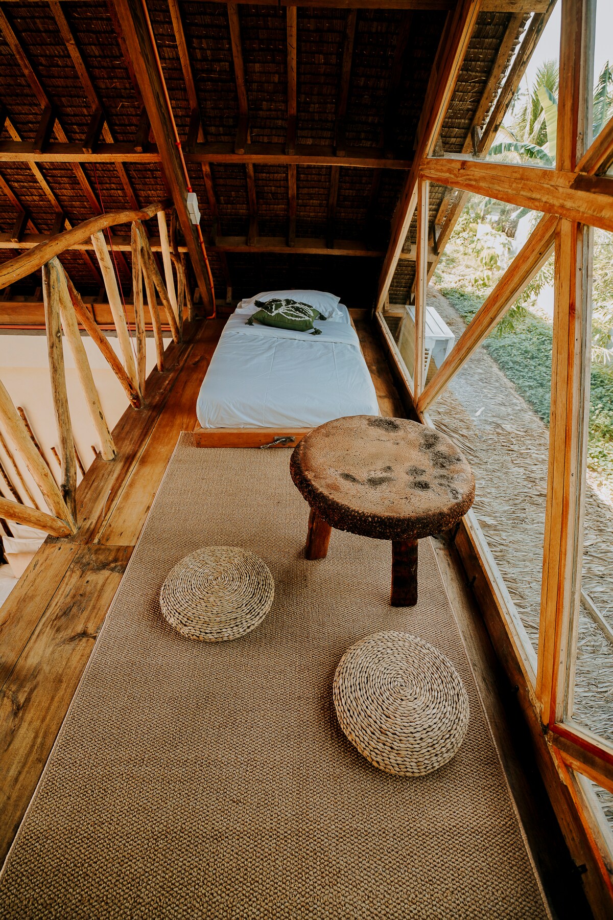 An open space on the second floor features a single bed and a small round table made from wood. Two woven stools sit on a natural fiber rug, with large windows allowing natural light to illuminate the room and offering views of the surrounding greenery.