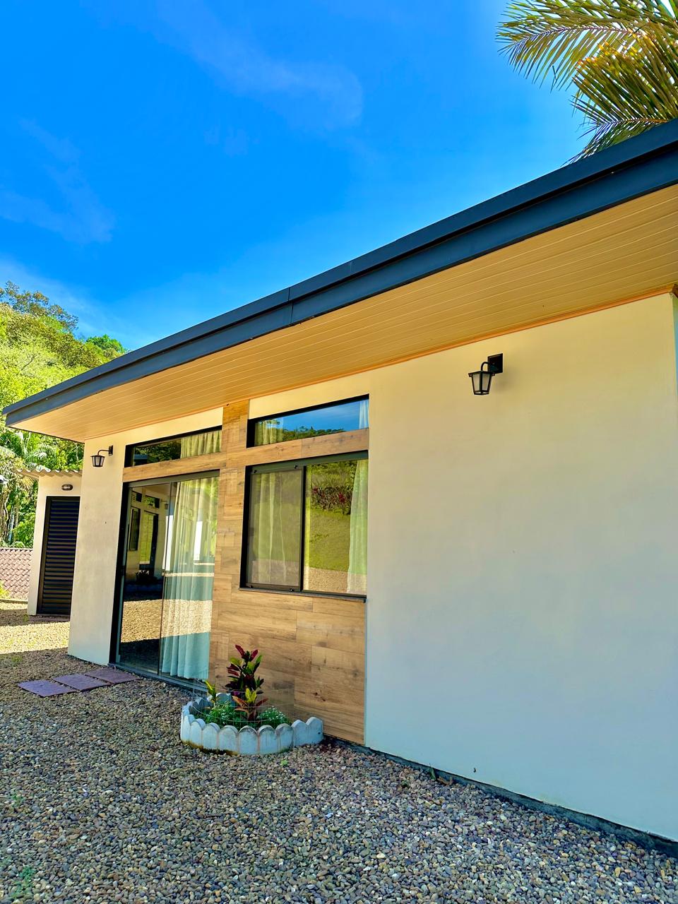 The exterior of the chalet showcases a modern design with a blend of white and wood accents. Large windows allow natural light to fill the interior, while a small garden area with decorative plants enhances the entrance. A clear blue sky complements the tranquil setting.