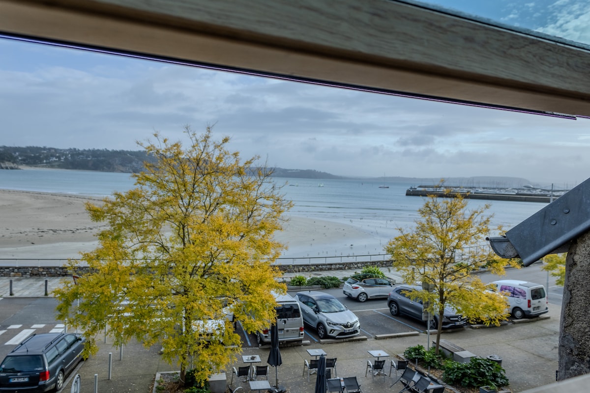 A view from a window reveals a serene beach landscape, with soft sand stretching toward the water. Autumn trees with yellow leaves frame the scene, while parked cars and a harbor are visible in the background.