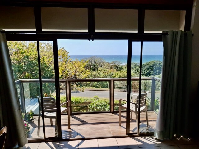 A sliding glass door opens to a private balcony, revealing a scenic view of the sea and lush treetops. Two outdoor chairs are positioned on the wooden deck, providing a comfortable space to enjoy the natural surroundings.