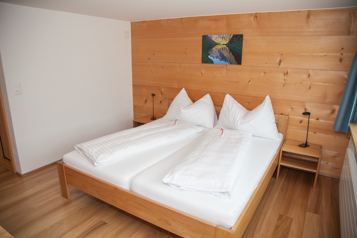 A bedroom features a spacious wooden bed with two peaked pillows and white bedding. Light wooden walls and floor create a warm environment. A small nightstand is positioned on one side, while a landscape artwork adorns the wall above the bed.