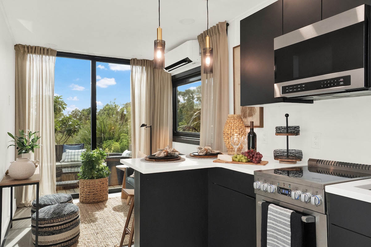 A modern kitchen is featured with sleek black cabinetry and stainless steel appliances. Soft drapes frame a large window, allowing natural light to brighten the space. A dining area with a woven rug includes decorative elements like plants and fruits, adding warmth to the overall design.