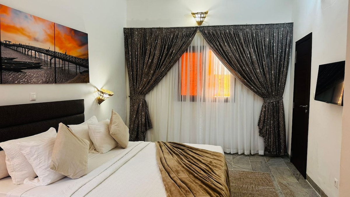 A cozy sleeping area features a spacious bed with plush pillows and a warm-toned blanket. Elegant drapery frames a window, allowing soft light to filter through. A vibrant artwork depicting a pier is displayed above the bed, adding a touch of coastal charm.