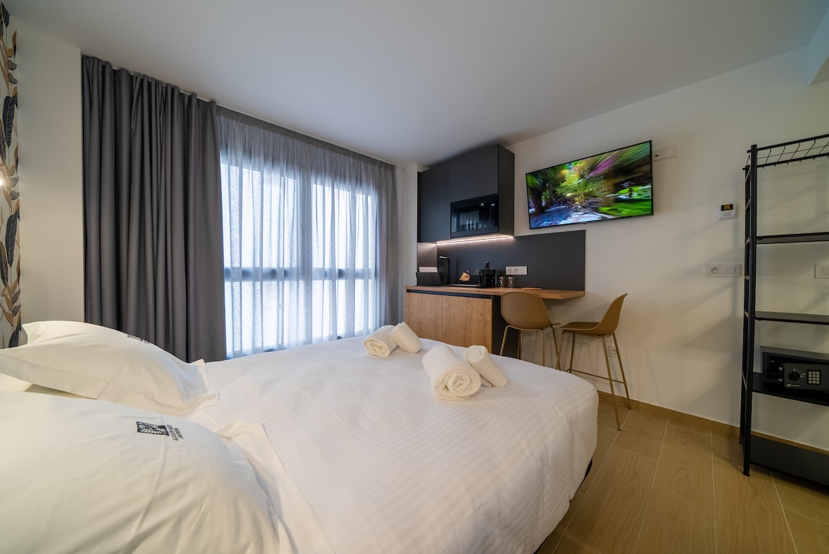 A well-lit sleeping area features a spacious bed with neatly rolled towels on top. Natural light filters through the sheer curtains covering large windows. A small workspace with a desk and two chairs is positioned nearby, complemented by a mounted television on the wall.