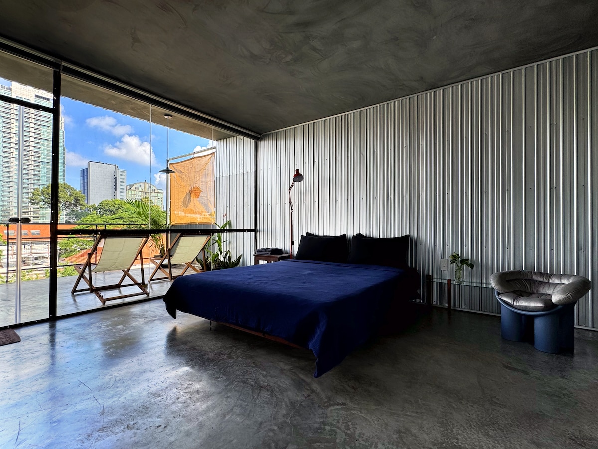 A spacious bedroom features a sleek design with a large bed dressed in dark linens. Natural light streams through expansive windows, illuminating the room. A small table and chair are positioned on the balcony, offering views of the surrounding cityscape and greenery.