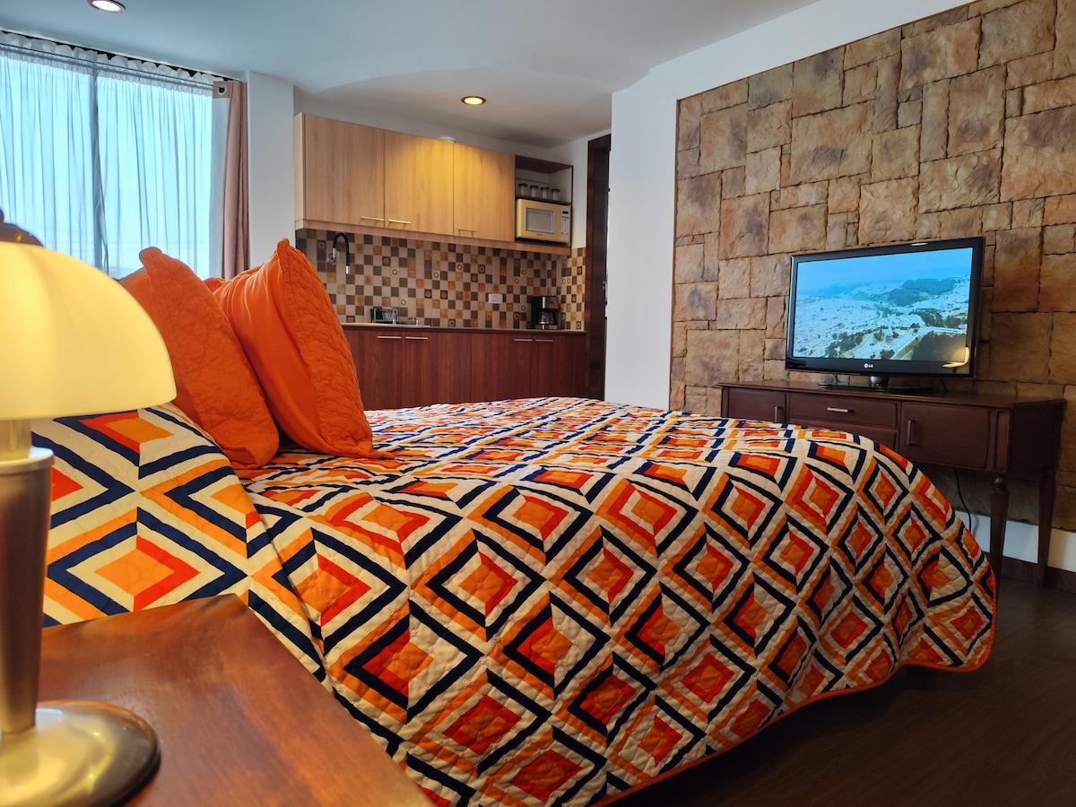 A spacious sleeping area features a brightly patterned blanket laid over a comfortable bed with orange and blue accents. A media console holds a television, while a kitchenette with wooden cabinetry is visible in the background. Natural light filters through sheer curtains.