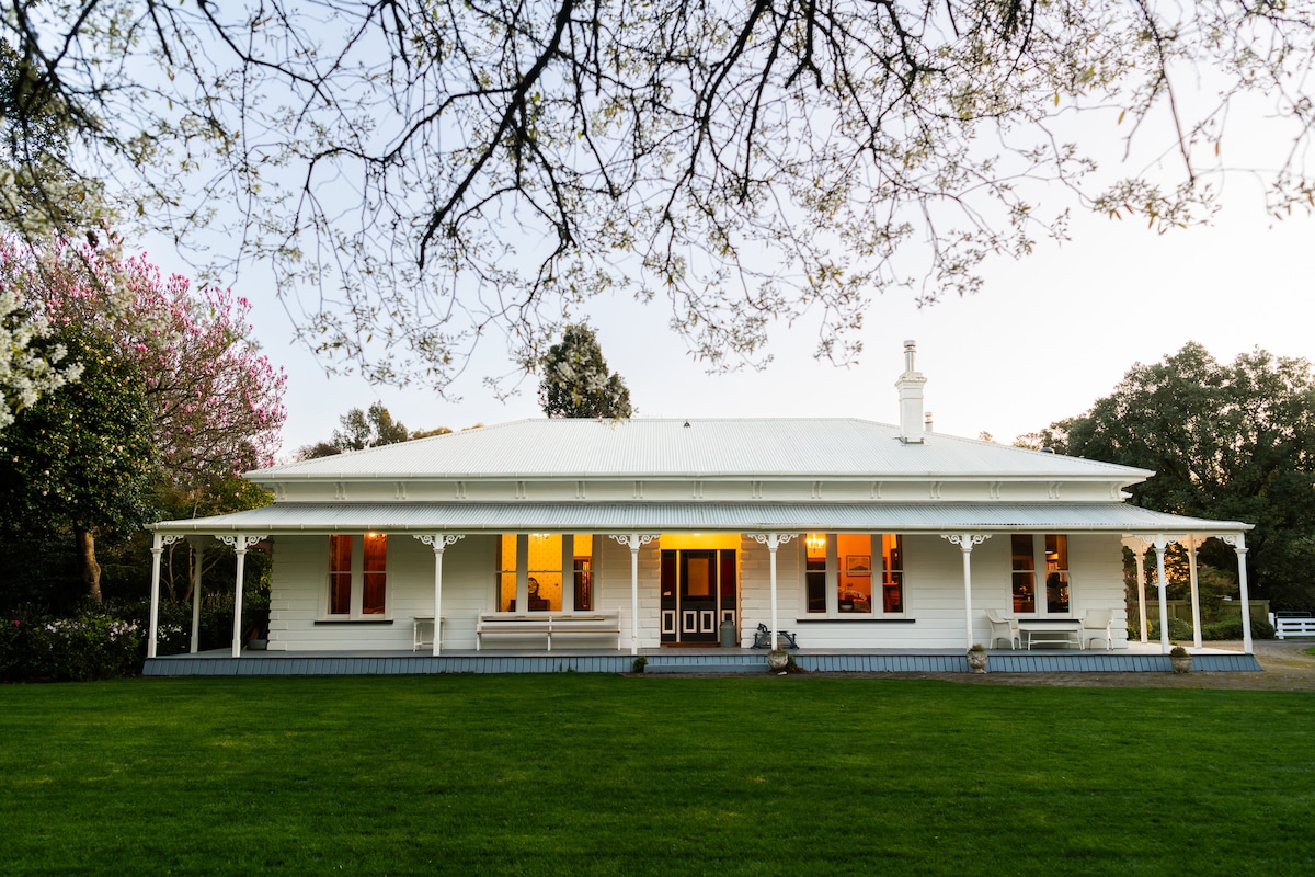 The historic farm homestead is showcased with a charming facade, featuring white painted wood and a welcoming front porch. Lush green grass surrounds the property, and colorful trees frame the scene, enhancing the serene atmosphere of the gardens.