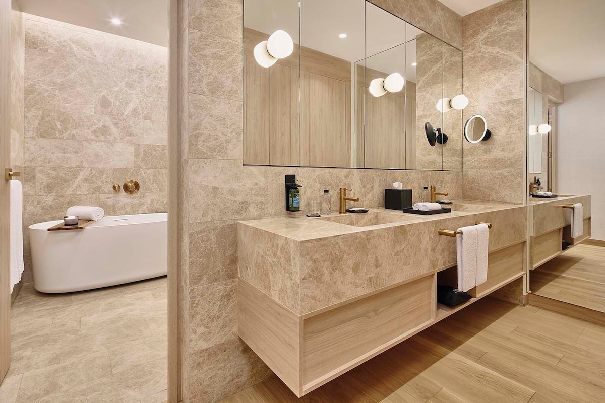 A spacious bathroom is designed with natural stone walls and a wooden vanity. Two mirrors are placed above the sink area, accompanied by stylish round lighting fixtures. A standalone bathtub is visible to the side, and elegantly folded towels are neatly positioned.