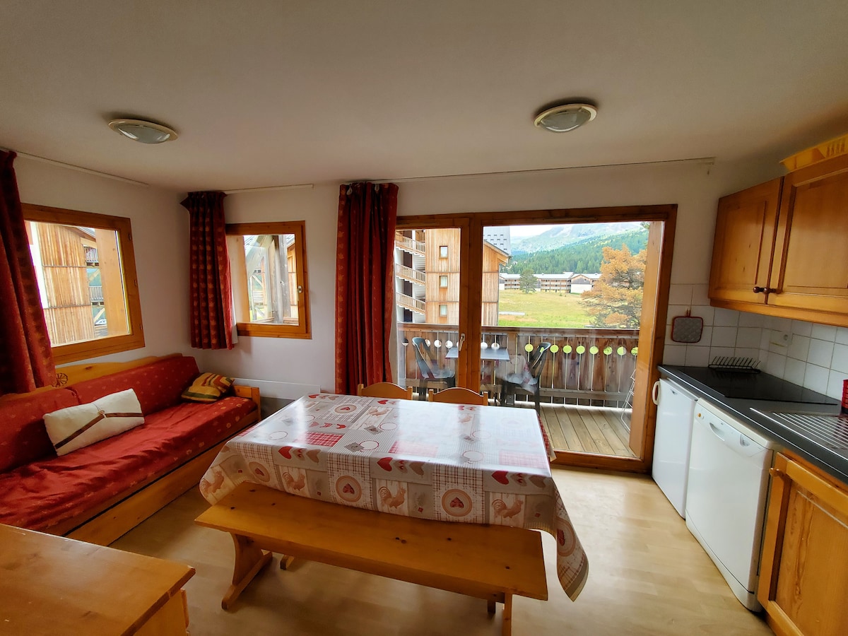 The spacious living area features large windows showcasing a view of the outdoors. A wooden dining table is surrounded by chairs. A cozy seating area is present with cushions, complemented by wooden cabinetry in the kitchen. Natural light brightens the room.