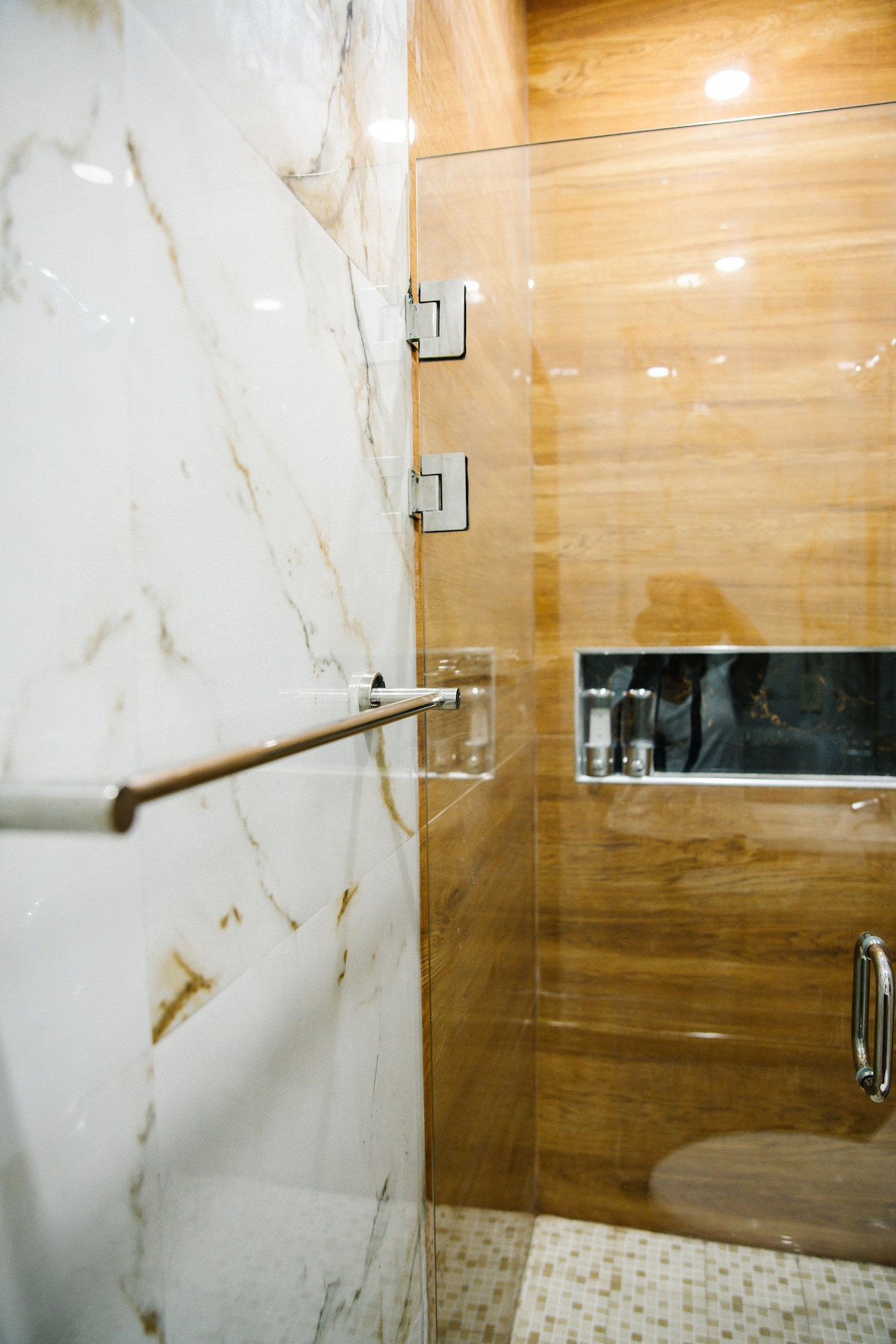 A modern shower area is enclosed by clear glass doors, revealing elegant tile work on the walls. A sleek grab bar is mounted for convenience, while warm wooden paneling adds a cozy touch to the space. Subtle lighting enhances the serene atmosphere.