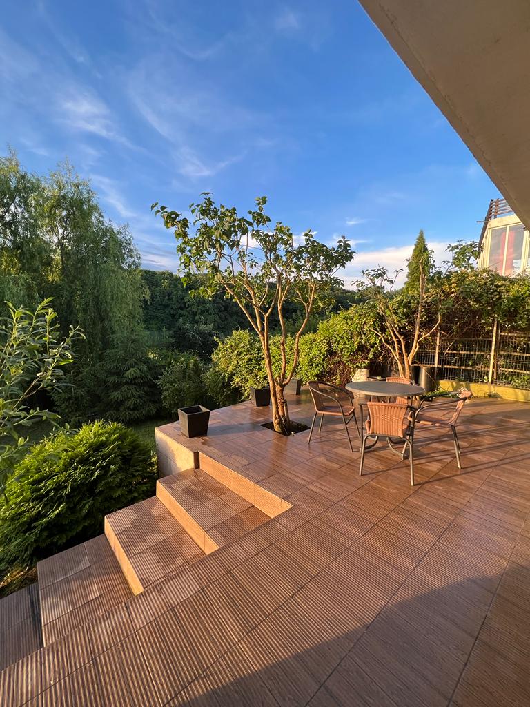 A spacious terrace area is presented, featuring a dining table surrounded by metal chairs. Lush greenery, including trees and shrubs, enhances the setting. Steps lead down from the terrace to a landscaped yard, framed by gentle slopes and a clear blue sky.