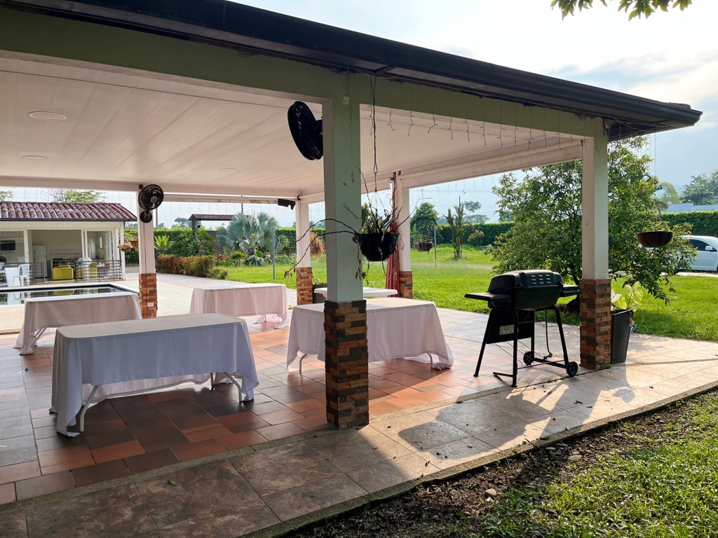 An outdoor dining area features several tables covered with white tablecloths, positioned on a tiled surface. A grill stands nearby, bordered by greenery. The space is shaded under a roof, providing a comfortable setting for gatherings.