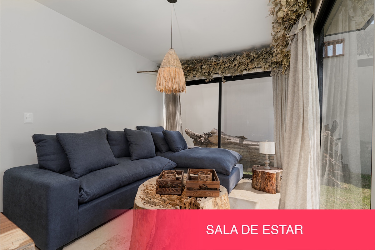 A comfortable seating area features a plush, dark blue sectional sofa, complemented by natural wood accents. A pendant light hangs overhead, while large glass doors offer a view of the outdoors. Textured curtains frame the space, creating an inviting atmosphere.