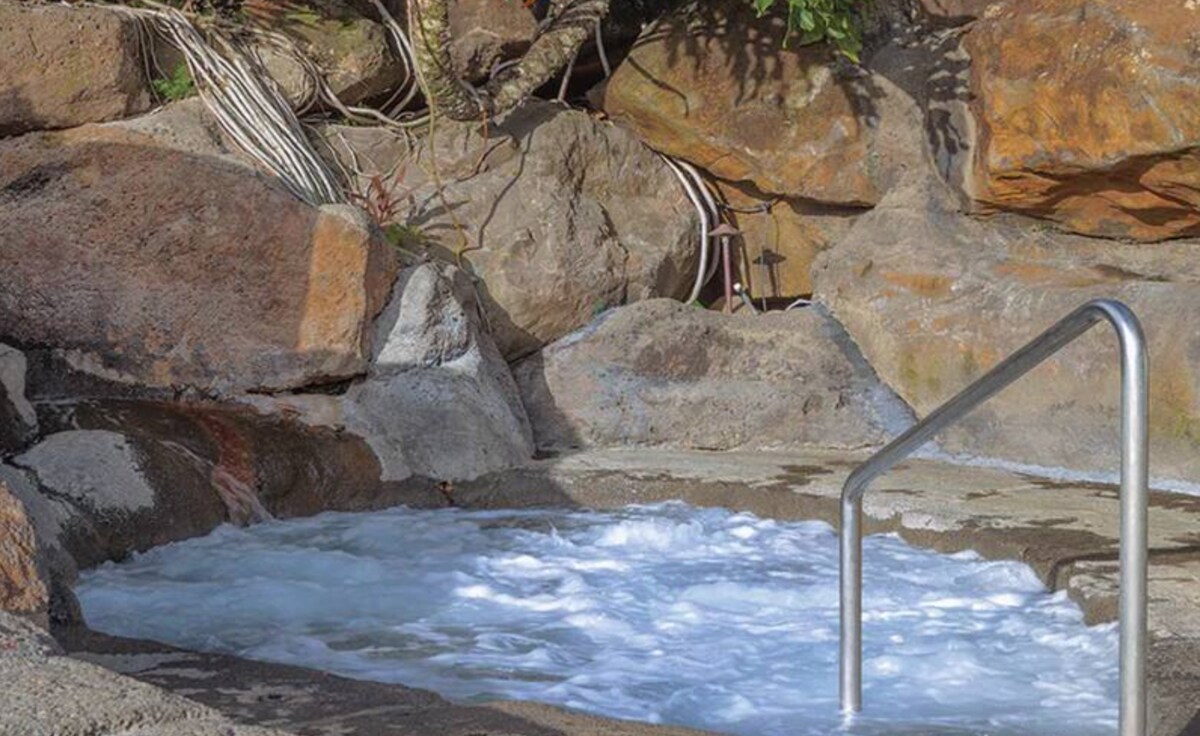 A bubbling jacuzzi is set against a backdrop of natural stone, highlighting the textured rocks and surrounding greenery. The water creates gentle waves, and a stainless steel railing provides support for entry. A tranquil setting invites relaxation and enjoyment of the surrounding nature.