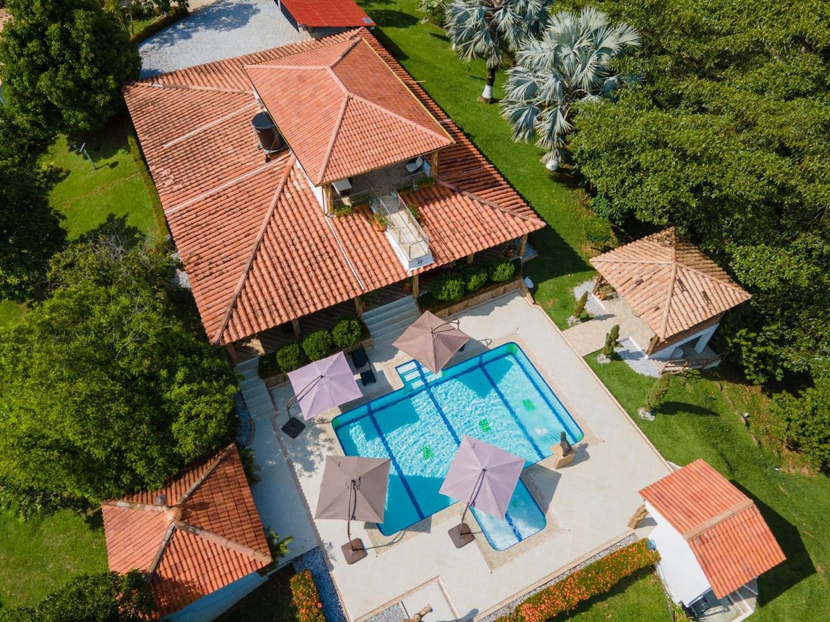An aerial view of the property reveals a spacious layout featuring a private swimming pool surrounded by umbrellas for shade. The house has a traditional tile roof and is complemented by lush green gardens and palms, adding to the serene outdoor ambiance.