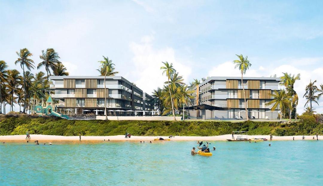 The modern resort features a contemporary architectural design, with expansive glass windows and multiple levels. Palm trees line the sandy beach, while guests engage in various water activities in the calm turquoise water. The lush green landscape enhances the serene coastal setting.