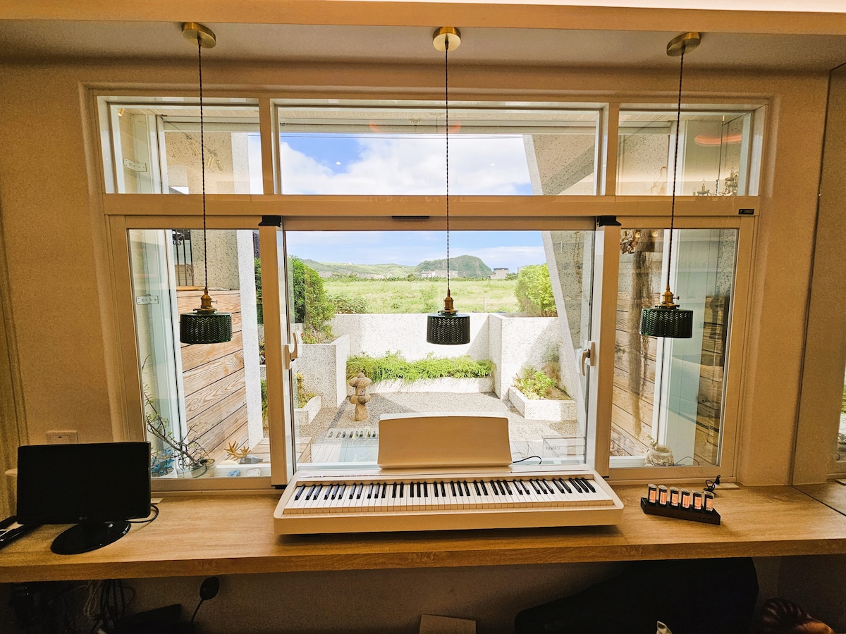 A window designed to frame a scenic outdoor view is adorned with two pendant lights. A keyboard rests on a wooden countertop, with a small black device positioned nearby. The garden outside features greenery and a stone pathway, offering a calm atmosphere.