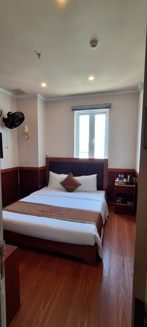 A well-lit bedroom features a spacious bed with a decorative throw pillow. Wooden accents create a cozy atmosphere. A small bedside table holds amenities, and a window allows natural light to enter while offering a glimpse of the outside.