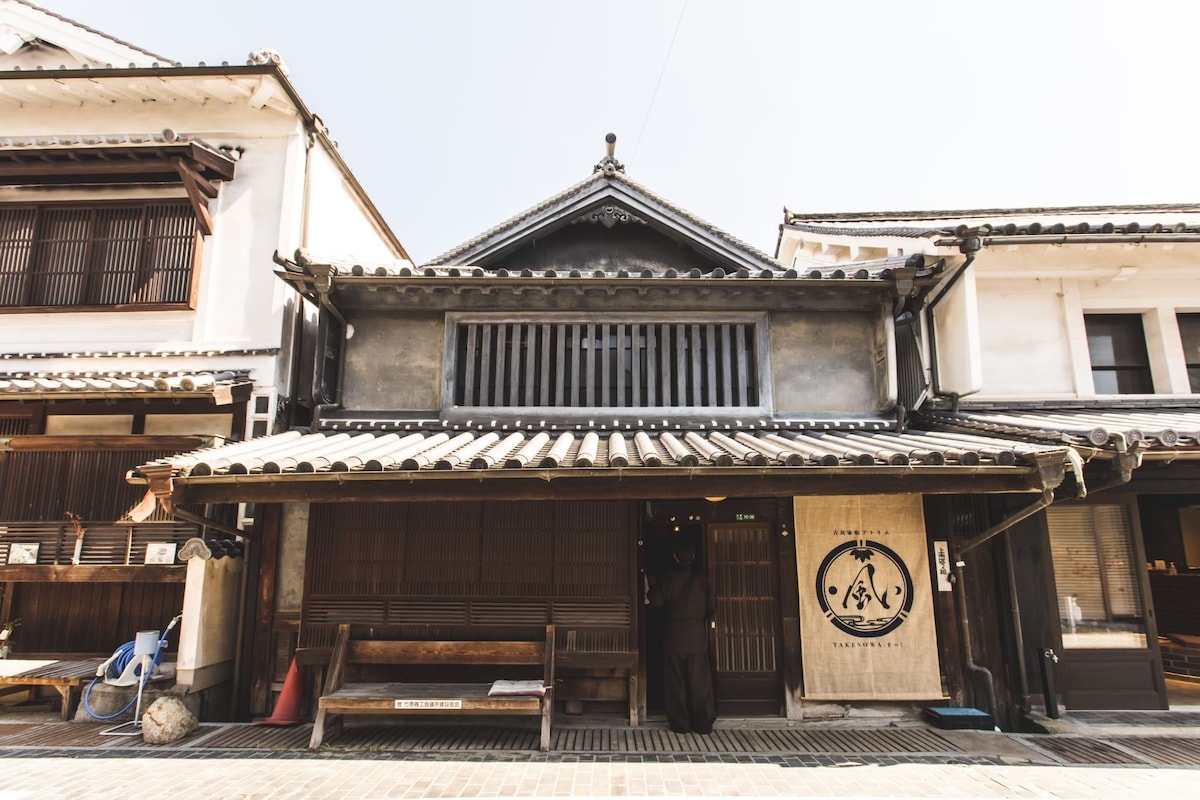 The exterior of a traditional two-story wooden house is depicted, showcasing its distinct architectural features. A tiled roof and wooden beams are prominent, with a wide entrance and a bench providing a welcoming touch. The building's historical significance is evident in its design.