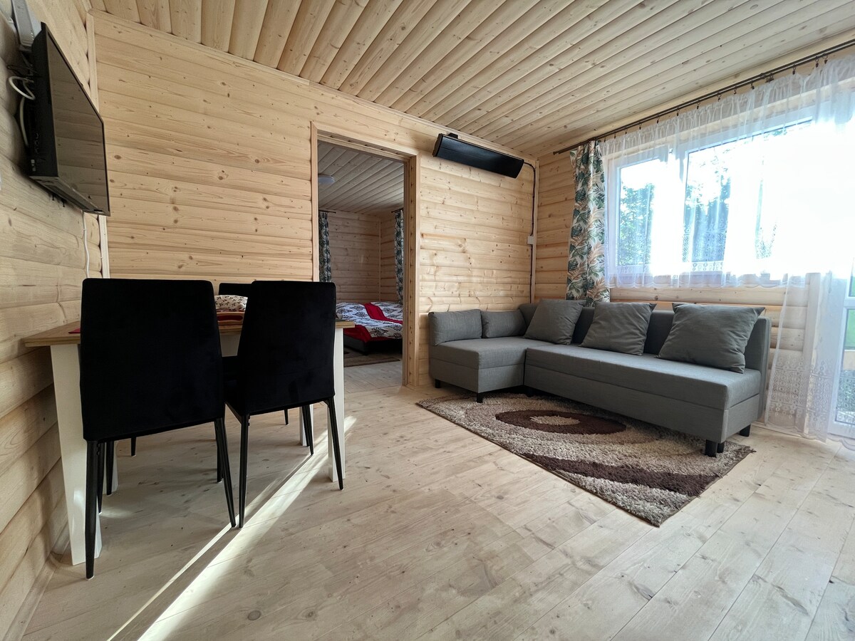 A light-filled living area features natural wood paneling and a cozy sectional sofa adorned with soft cushions. A dining table with four chairs is positioned nearby, while a large window offers views of the serene outdoor landscape and lets in ample sunlight.