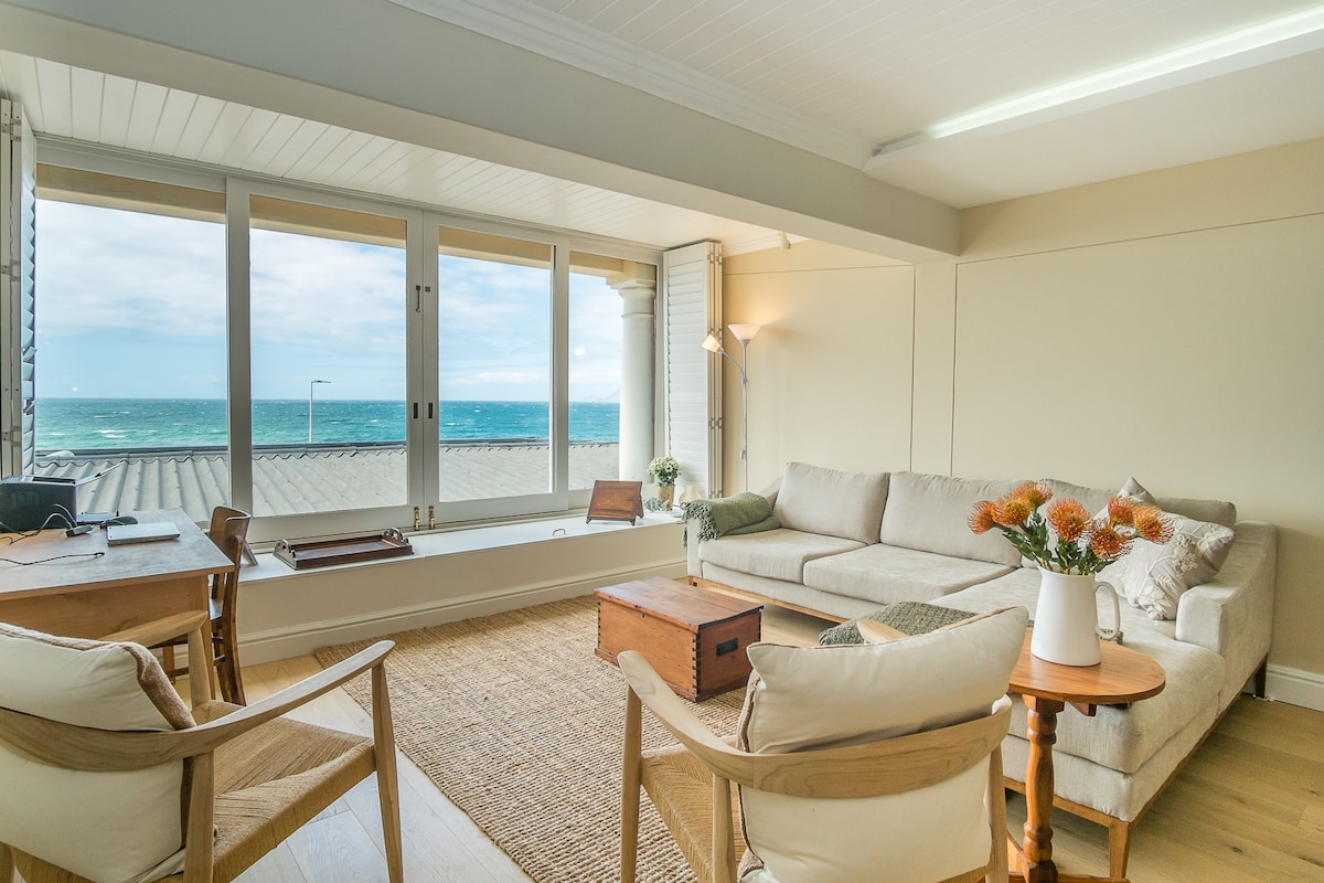 The living area features large windows offering unobstructed views of the sea. A comfortable sofa is positioned beside a wooden table. Two minimalist chairs are thoughtfully placed, and a small table holds a vase with flowers, enhancing the space's tranquil atmosphere.