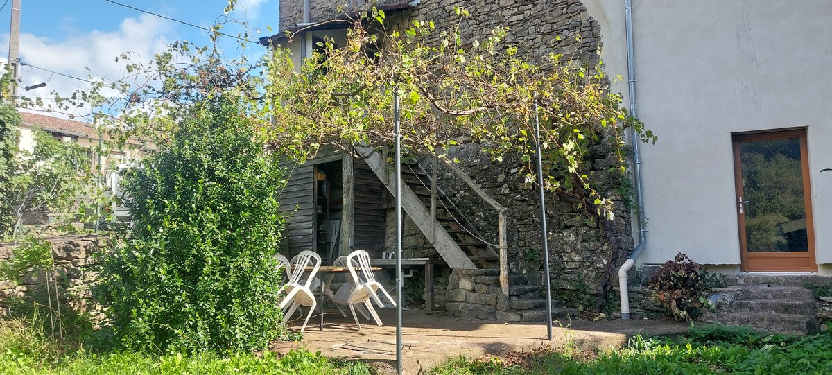 An outdoor area is featured, showcasing stone walls and a staircase leading to an entrance. Two white chairs are placed around a small table, and lush greenery surrounds the space, including vines overhead. Sunlight filters through the trees, creating dappled light on the ground.