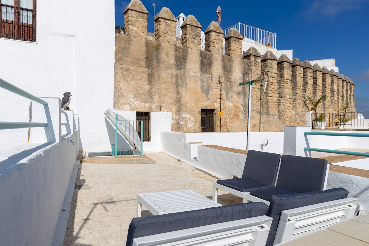 An outdoor terrace features comfortable seating with two lounge chairs and a small table, overlooking a historic stone wall. The wall displays rounded crenellations, providing a glimpse of textured masonry and architectural detail, with clear blue skies enhancing the view.