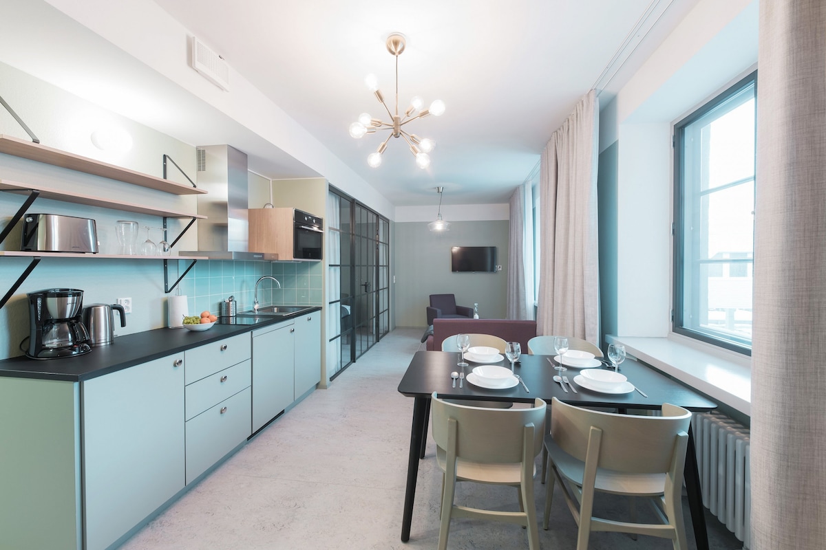 The kitchen area is equipped with modern appliances, including a coffee maker and oven, complemented by a sleek dining table set for four. Large windows allow natural light to brighten the space, while a cozy seating area is visible in the background.