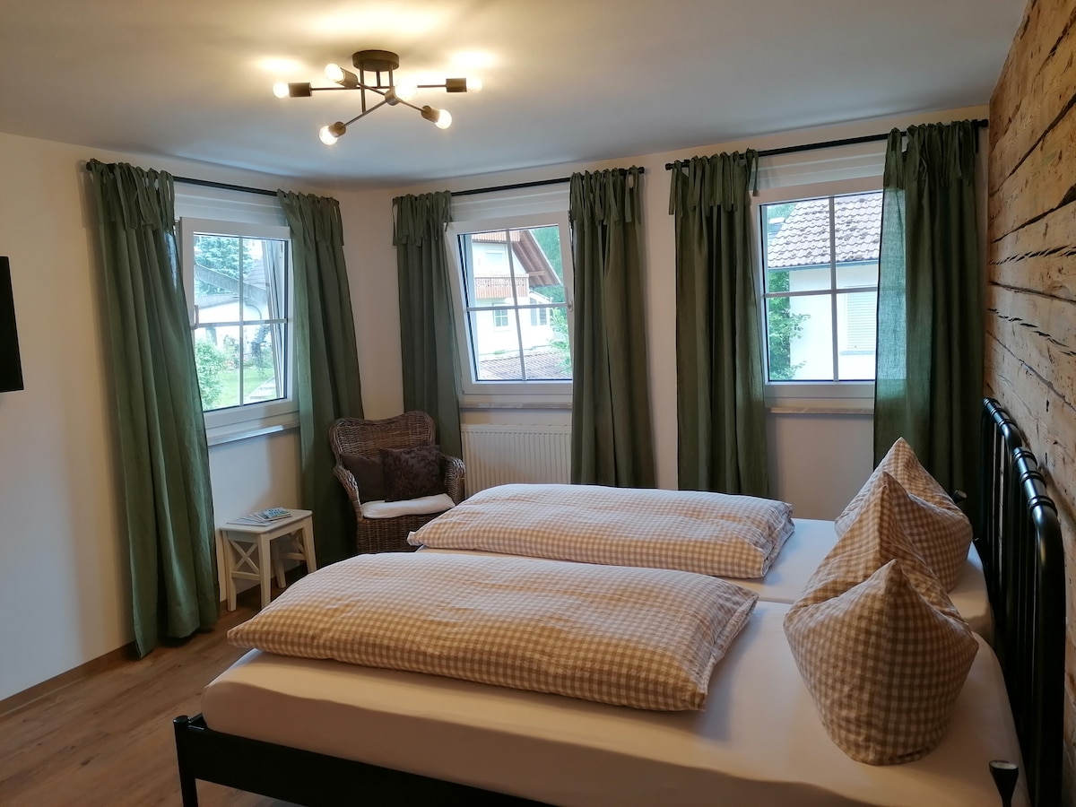 A well-appointed bedroom is presented with a spacious bed featuring two separate mattresses and decorative pillows. Natural light enters through two windows dressed in green curtains. A comfortable armchair is placed beside a small side table, enhancing the room's welcoming ambience.