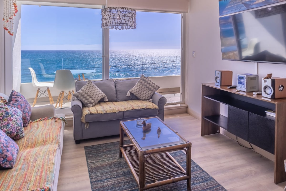 A bright living area features a comfortable sofa adorned with colorful cushions, while a glass coffee table sits on a textured rug. Large windows provide a view of the ocean, allowing natural light to fill the space. A media console holds entertainment equipment.