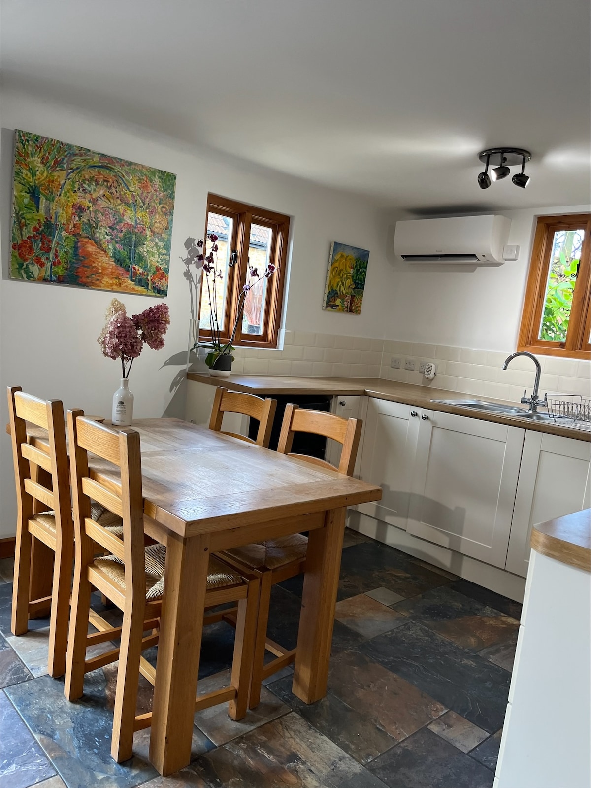 A well-equipped kitchen is depicted, featuring a wooden dining table with four chairs. Bright artwork adorns the walls, and natural light filters through the windows. A modern air conditioning unit is mounted on the ceiling, enhancing comfort.