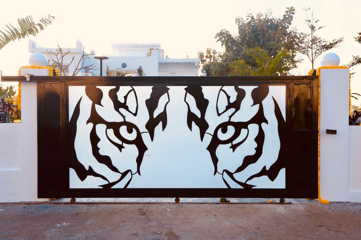 A striking entrance gate features a bold design of tiger eyes in black and white. The artistic gates are framed by white walls and complemented by lush greenery in the background, setting a unique and inviting tone for the property.