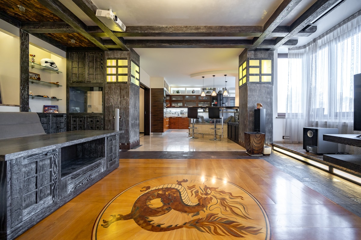 An open living space features a finely crafted wooden floor with an intricate dragon design at its center. The room boasts wooden accents and large windows that allow natural light to fill the area. A modern kitchen is visible in the background, adding to the space's functionality.