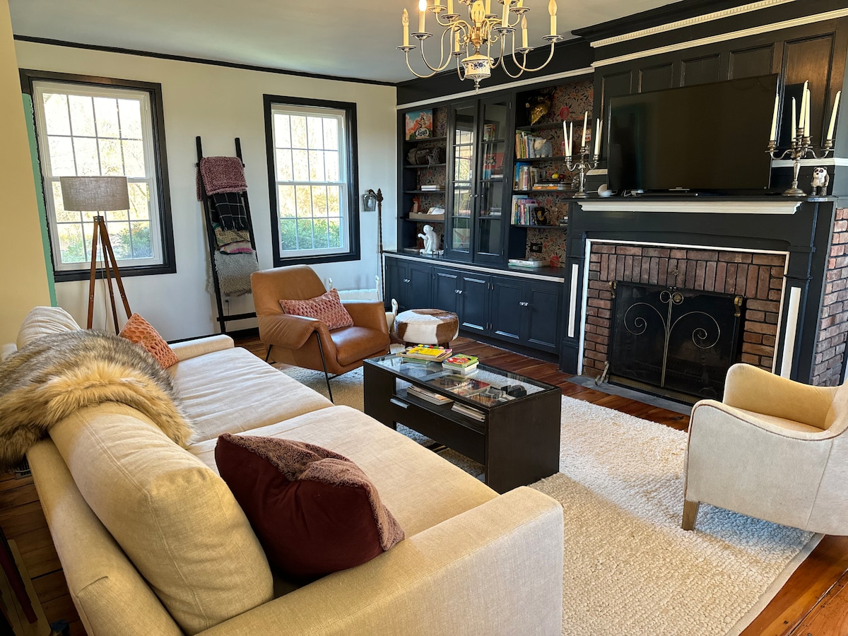 A spacious living room features an extra deep sofa and two comfortable armchairs. A television is mounted above a brick fireplace, with a bookcase displaying various items. Natural light filters through the large windows, creating a bright and inviting atmosphere.