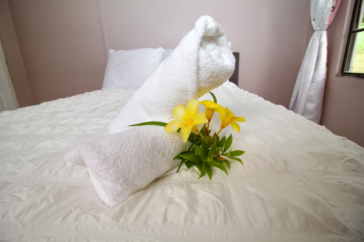 Two rolled white towels are placed on a neatly made bed, accompanied by a small arrangement of yellow flowers and greenery. The neutral-colored walls and soft linens create a serene setting, enhancing the inviting look of the sleeping area.