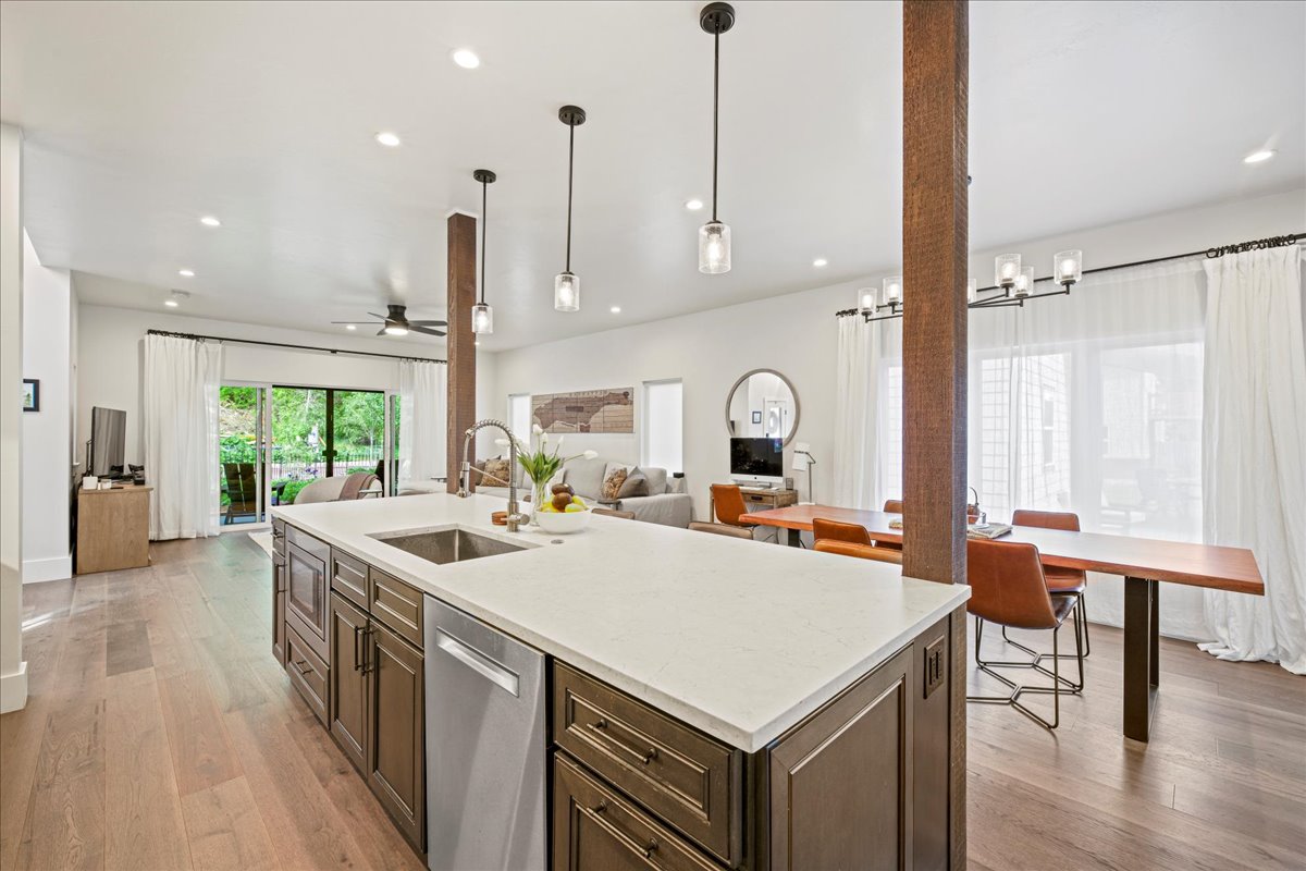 An open-concept kitchen and dining area features modern cabinetry and a large island with a sink. Bright overhead lighting complements the warm wood accents. Floor-to-ceiling windows provide natural light and views of the outside space. A dining table is positioned nearby, accompanied by stylish chairs.
