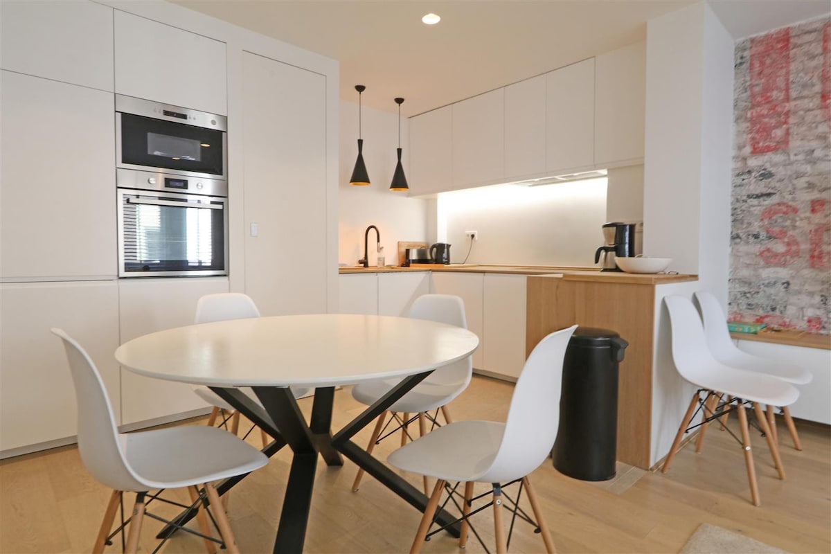 A bright and modern kitchen space features sleek cabinetry and stainless steel appliances. A round dining table, surrounded by four white chairs, provides a cozy area for meals. The minimalist design is complemented by contemporary lighting fixtures above the countertop.