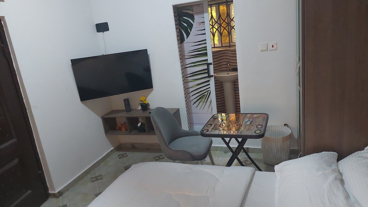 A cozy studio room is depicted with a spacious bed featured prominently. A small table with a decorative design stands next to a comfortable chair. A wall-mounted television is visible, and a door with decorative glass leads to a light-filled outdoor area.