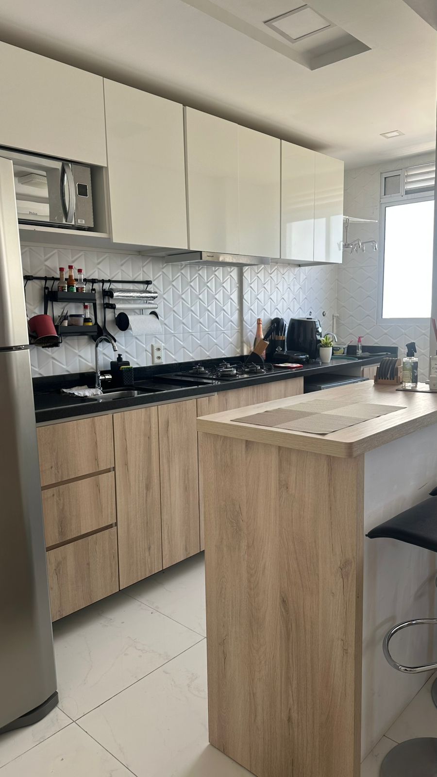 A modern kitchen is presented, featuring sleek cabinetry in white and wood tones. A stainless steel refrigerator stands beside a countertop with a stovetop and various cooking utensils. Natural light enters through a nearby window, illuminating the clean, functional layout.