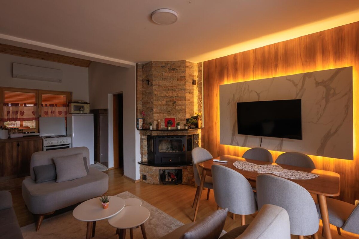 A welcoming living area is highlighted by warm lighting and a modern design. A fireplace is positioned in front of a wall-mounted television. A dining table with chairs is placed beside a cozy seating arrangement, enhancing the inviting atmosphere of the space.