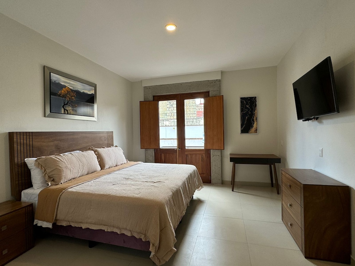 A spacious bedroom features a large bed with neutral-toned bedding set against a light wall. Natural light enters through the wooden windows, complemented by a modern artwork hanging above the bed. A wooden desk and dresser provide functional storage solutions.