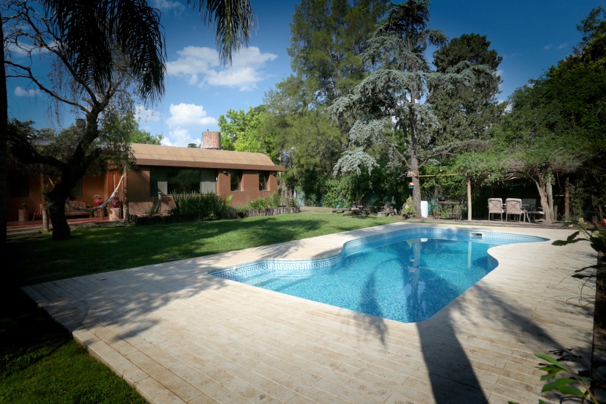 A private pool is framed by a spacious deck, surrounded by lush greenery. In the background, a charming house features a warm-toned facade and a thatched roof. Soft sunlight casts gentle shadows across the outdoor space, enhancing the tranquil atmosphere.