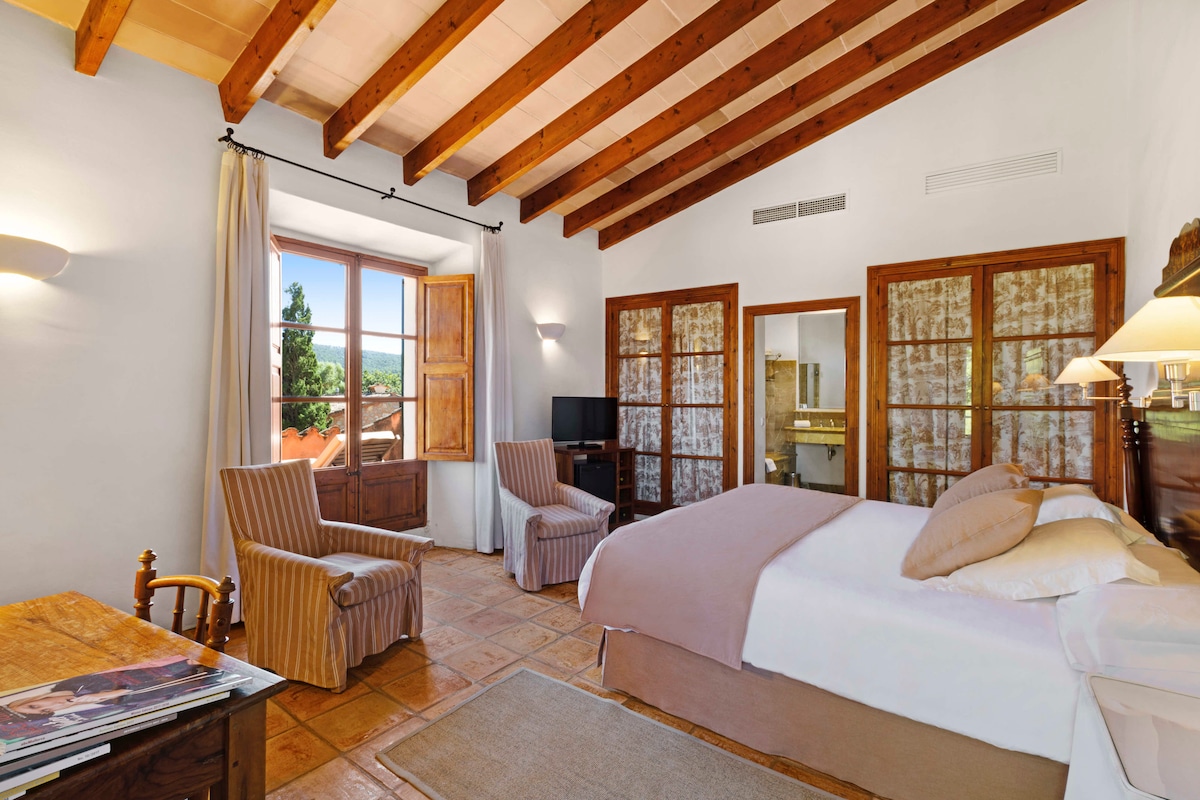 The room features exposed wooden beams and terracotta tiles. A spacious bed is complemented by bedside lamps. Two armchairs and a wooden desk create a cozy seating area. A large window allows natural light to illuminate the space, revealing views of the surrounding greenery.