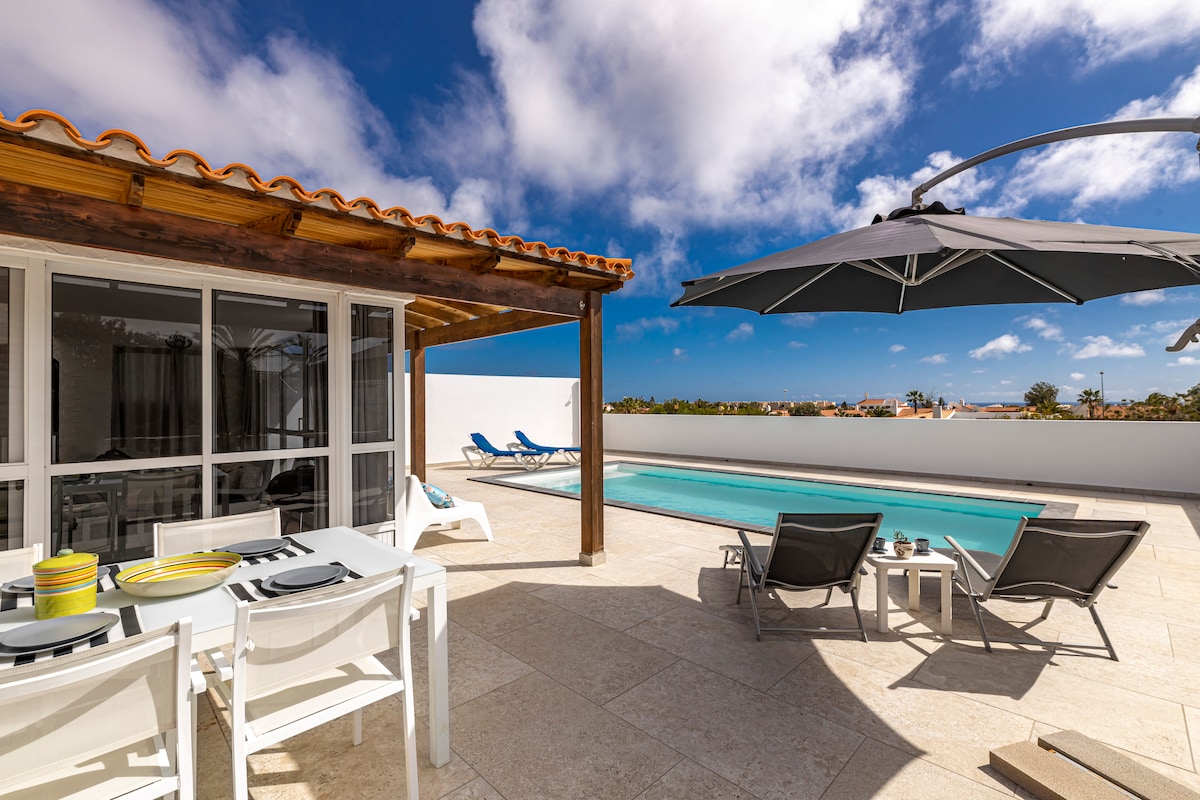 The outdoor terrace features a private swimming pool surrounded by lounge chairs. A shaded dining area with a table and tableware overlooks the pool, and a large umbrella provides additional shade. The backdrop includes clear skies and views of the surrounding area.